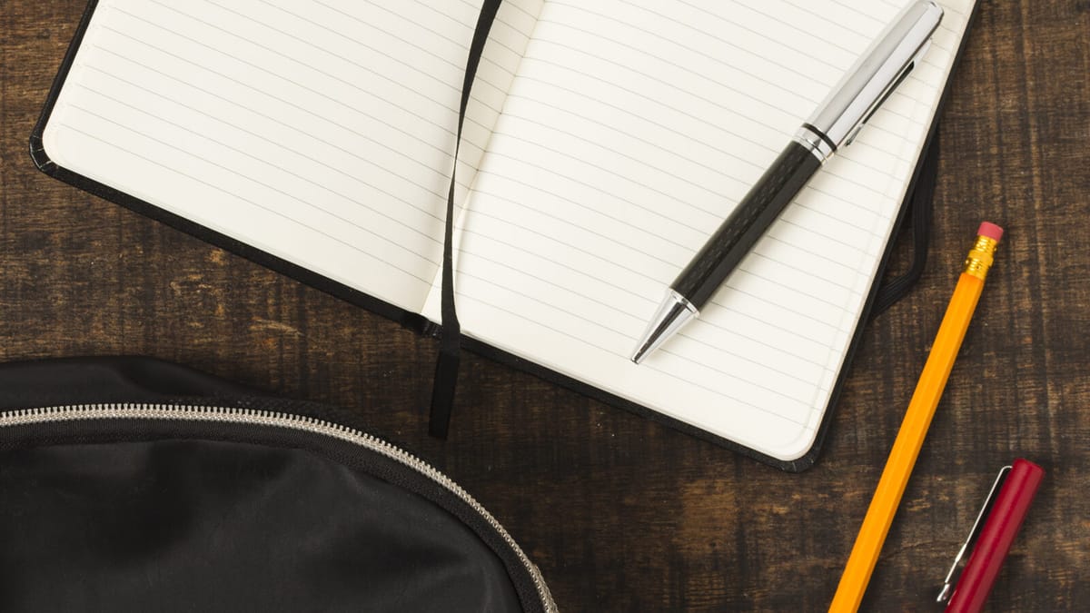 Caderno aberto, caneta, lápis e mochila sobre uma mesa de madeira