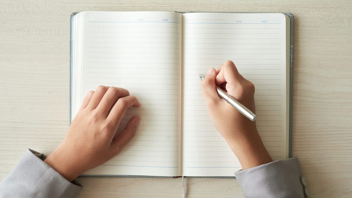 Caderno pautado aberto sobre a mesa, com as mãos de uma pessoa segurando a caneta