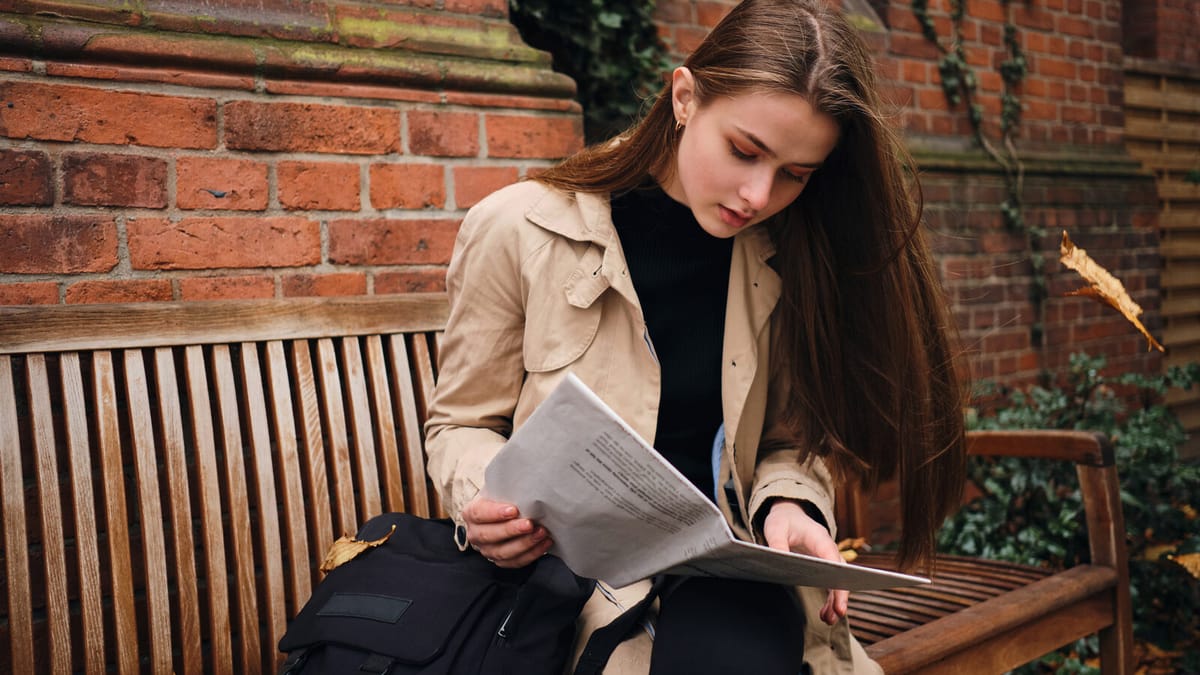 Jovem lendo um jornal sentada em um banco, com a mochila ao lado
