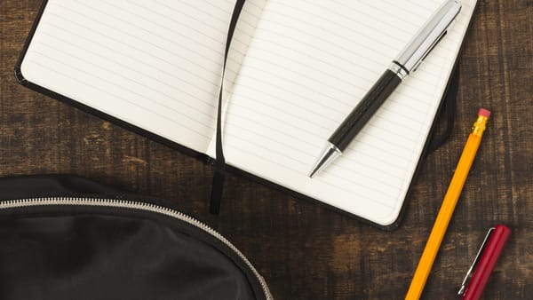 Caderno aberto, caneta, lápis e mochila sobre uma mesa de madeira