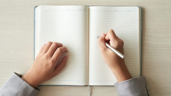 Caderno pautado aberto sobre a mesa, com as mãos de uma pessoa segurando a caneta